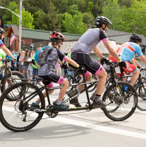 Cyclists race on a sunlit gravel road surrounded by lush green trees.
