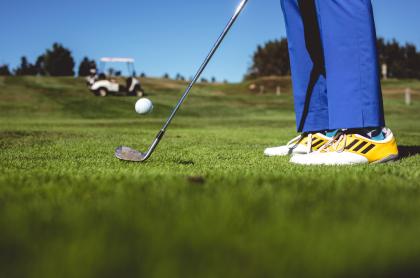 picture from the golf green of person in yellow and white golf shoes and bright blue pants&#44;hitting a golf ball with a pitching wedge. there is a golf cart on a hill in the backgroung