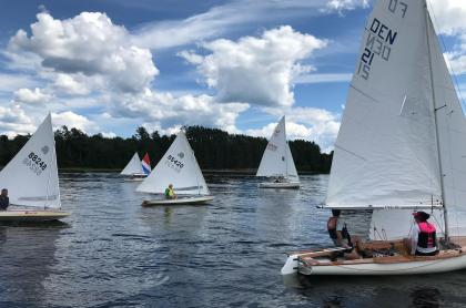 5 small sail boats with white sails on the lake with the mountains and trees in the background and a blue sky full of white puffy clouds