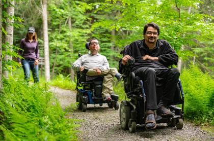 Three people heading down a trail in the woods&#44; two of them are in wheelchairs