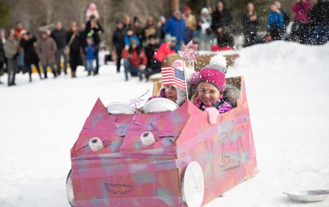 Two young girls ride in a cardboard sled in a race.