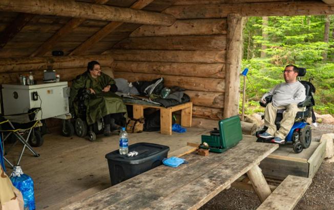 Two men in mobility chairs sit under a lean-to.