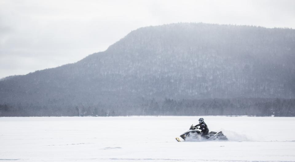 A snowmobiler riding across a snowy lake with mountain views in the background.