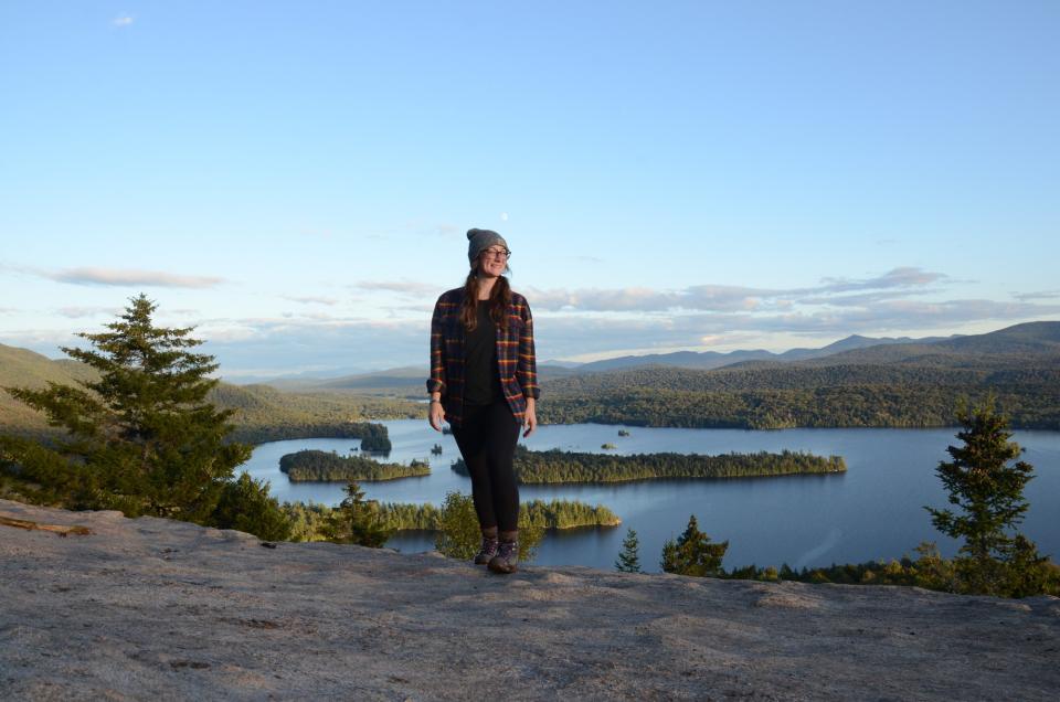 A woman walks along a mountain top with lakes in the background.