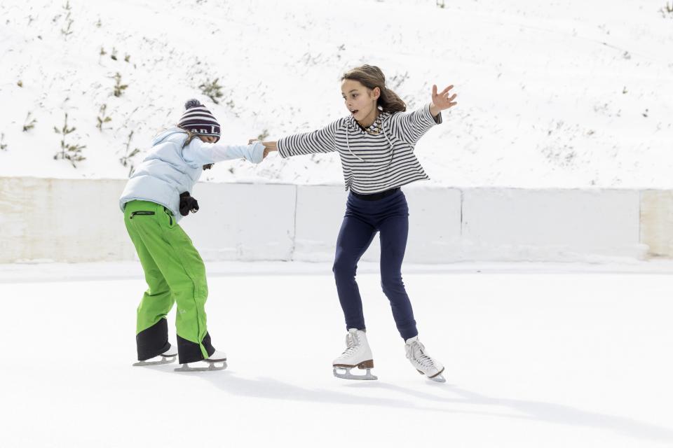 Two young girls ice skate on an ice skating rink.