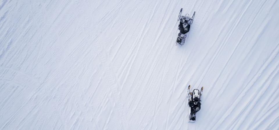 An aerial view of two snowmobilers riding across the snow.