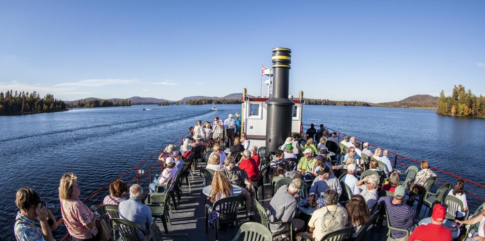 People mill around the upper level of a replica steamboat on an expansive lake with forested shoreline..