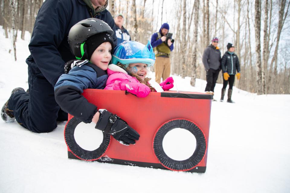 Two small children wearing winter clothing sit in a red sled made out of a cardboard box.