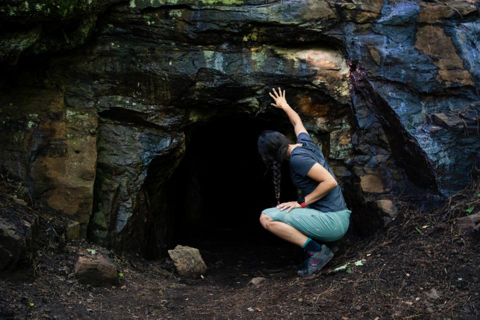 A young woman peers into the dark opening of a rock cave.