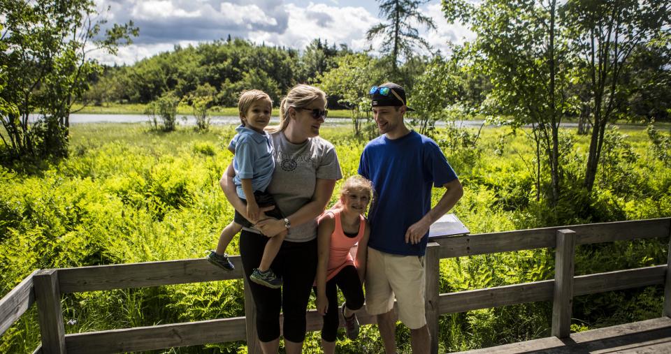 A man, woman, and two small children pose on a wood deck overlooking a wooded wetland.