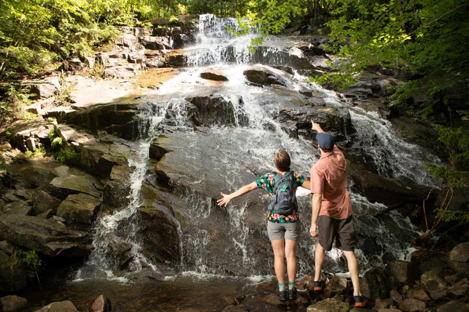 Two hikers look up in awe at a waterfall with green leaves on the side