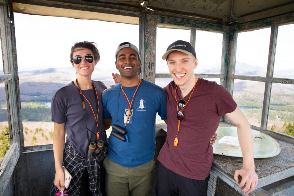 A young woman and two young men, posing for the photo in a fire tower over looking the Adirondack lakes and mountains in the background.