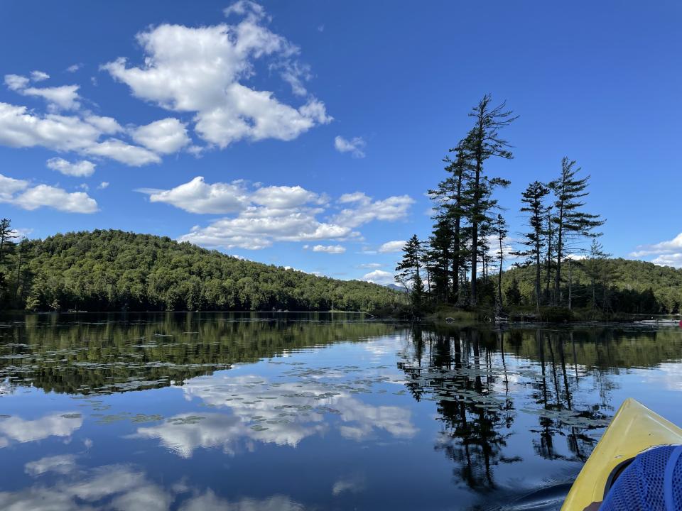 A view of Mason Lake from a yellow kayak with hills and pine trees in the background under a blue sky with some white puffy clouds, all reflecting into the water.