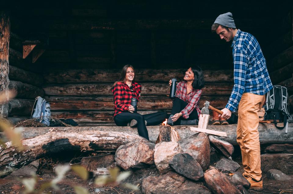 Two women in flannels laugh and drink from thermos's in a lean-to while a man stocks the fire.