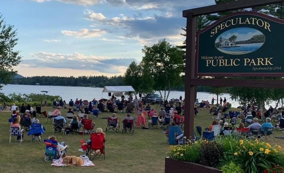 The sign of Lake Pleasant Public Park with many people sitting in camp chairs or on the ground listening to a band play at the Point with the view of Lake Pleasant in the background