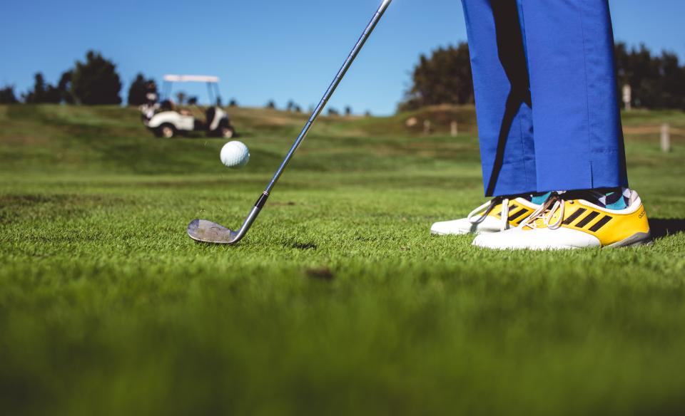 picture from the golf green of person in yellow and white golf shoes and bright blue pants,hitting a golf ball with a pitching wedge. there is a golf cart on a hill in the backgroung