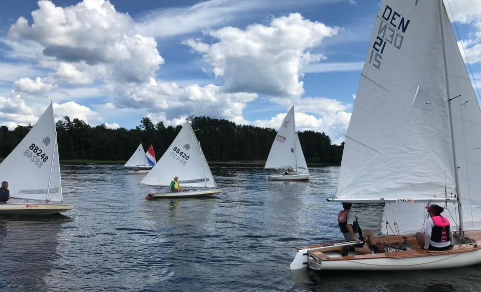 5 small sail boats with white sails on the lake with the mountains and trees in the background and a blue sky full of white puffy clouds