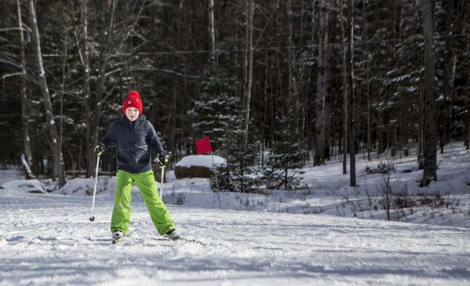 A boy with a red winter hat, black winter coat, and lime green snowpants, is cross-country skiing