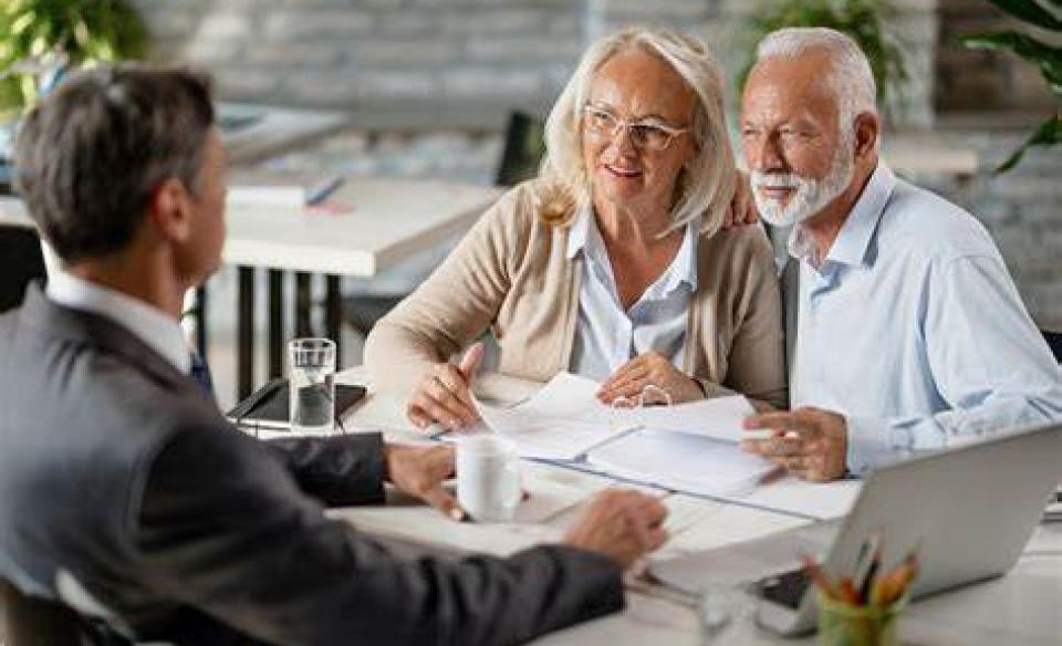 An elderly couple sitting at a table with paperwork in front of them with a man in a suit across the table