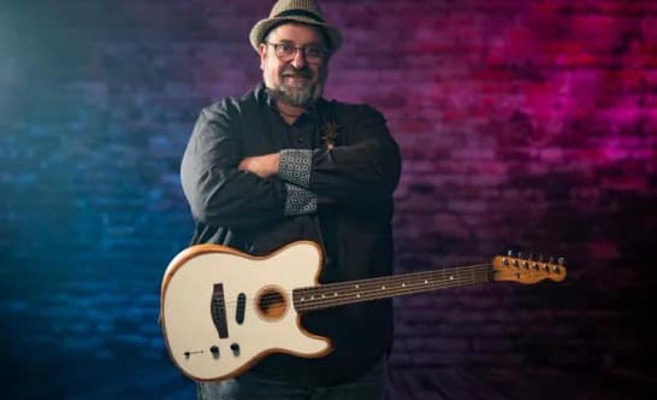 A photo of Eric standing with his arms crossed with his guitar in front of him.