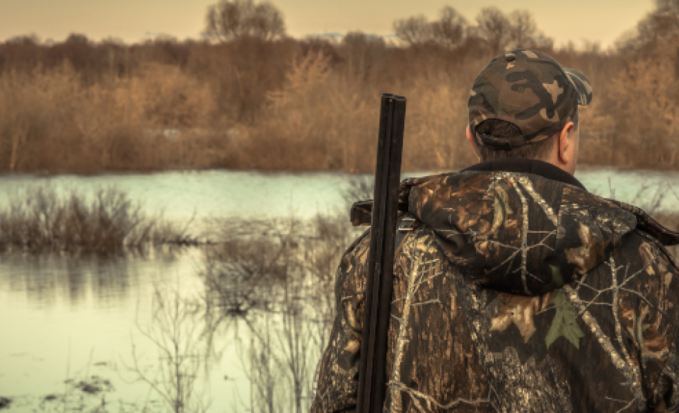 A man dressed in camouflage with a shotgun over his shoulder standing and looking at wetlands area